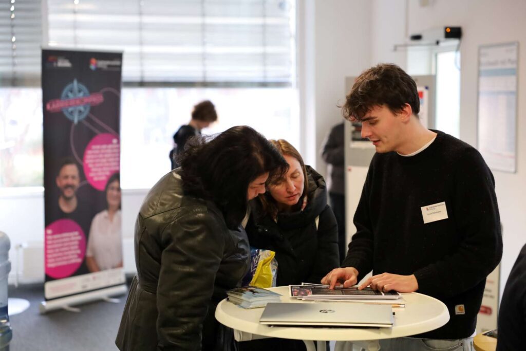 Ein Mitarbeiter am Infostand berät zwei Besucherinnen auf dem KarriereKompass 2. Titelbild für den Beitrag "Weiterbildungsbörse 2. KarriereKompass Südniedersachsen“ zum zweiten Mal angeboten."